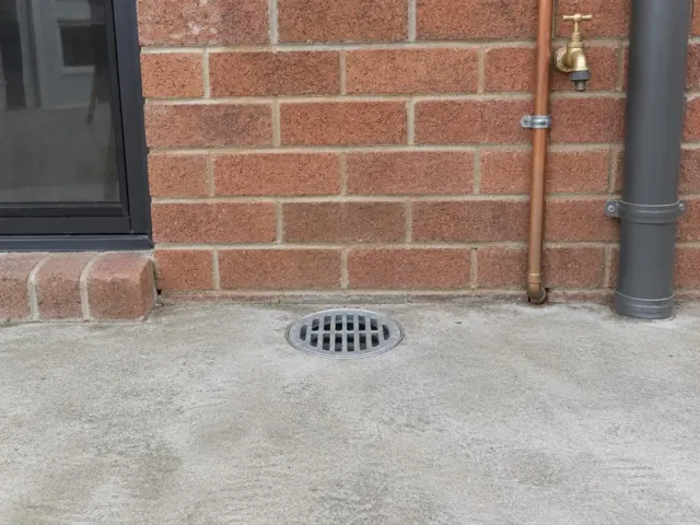 Round gully grate on dry concrete beside a brick wall and downpipe in Kellyville