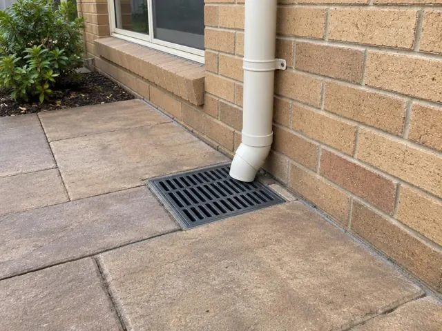 Seated stormwater grate beside a brick house wall and downpipe on dry paving in Bella Vista